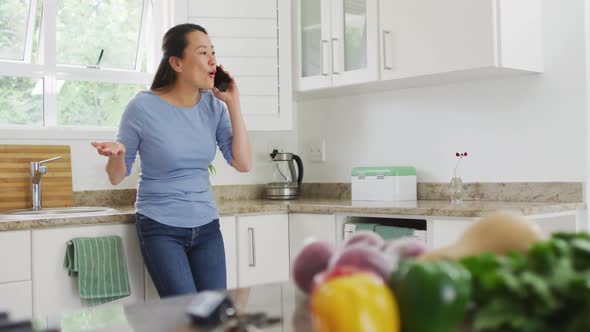 Happy asian woman standing at countertop and using smartphone in kitchen