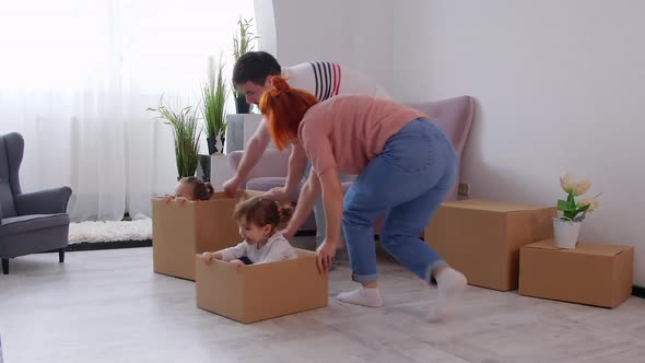 Excited happy young parents run push cardboard boxes with little cute childrendaughters ride inside