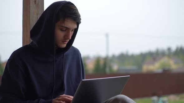 A Young Man is Typing on a Laptop While Sitting on a Terrace Against the Backdrop of a Picturesque