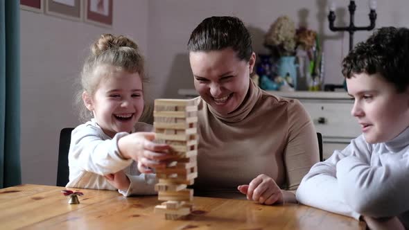 playful mom helps preschool children build tower of wooden blocks in living room