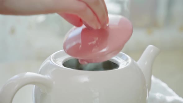 A woman's hand gracefully closes the lid of a white ceramic teapot with hot tea. Close-up.