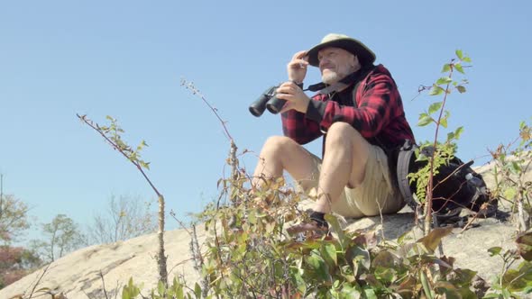 An older hiker looking through binoculars while on a scenic hike in the mountains.