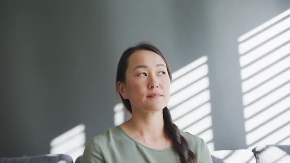 Thoughtful asian woman sitting on sofa close to window at home