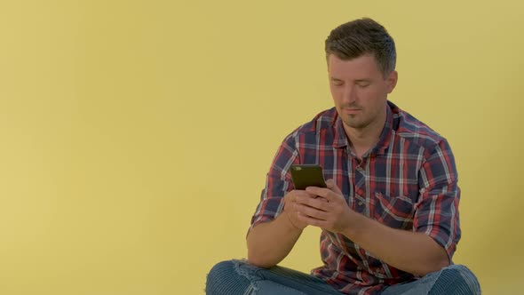 Cheerful Man in Checkered Shirt Sitting on the Floor and Chatting on Smartphone