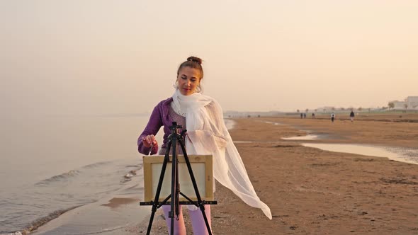 Delighted Artist Stands at Metal Easel Drawing on Beach