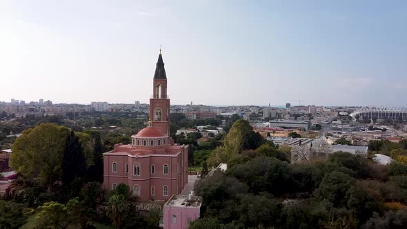 Aerial forward shot of a  pink church surrounded by trees, in front of city buildings and blue sky,