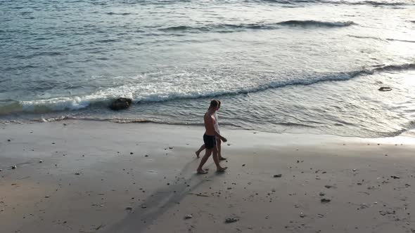 Man and Woman Walking Along Tropical Beach at Sunset, Tracking Aerial Shot of Young Couple on