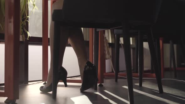 Close-up of Female Caucasian Legs in Elegant Black Shoes Sitting at Chair. Young Office Worker at