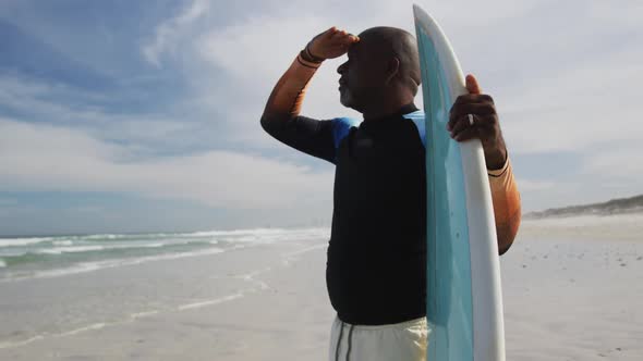 African american senior man standing on a beach holding surfboard and looking out to sea