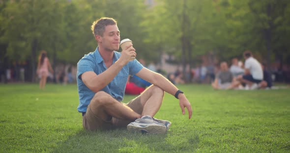 Portrait of Young Caucasian Man Drinking Takeaway Coffee Sitting on Lawn in Summer Park