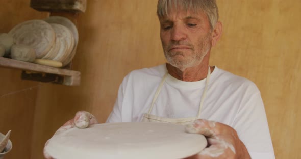 Senior caucasian man wearing apron inspecting clay dish in pottery workshop