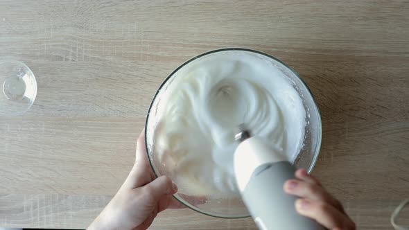 Female hands beating egg whites cream with mixer in the bowl on wooden table.