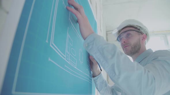 Young engineer-architect in a bright room in a helmet and glasses works with blueprints  alt