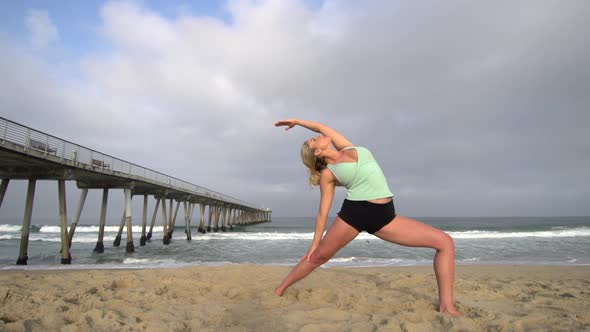 A young attractive woman doing yoga on the beach next to a pier.