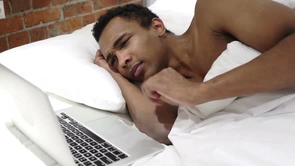 Pensive African Man in Bed Working on Project on Laptop