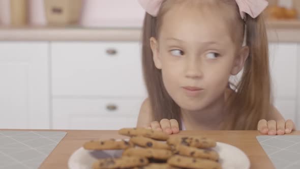 Cheerful Little Girl Peeking Out of Table and Taking Sweet Tasty Cookie From Plate, Portrait 