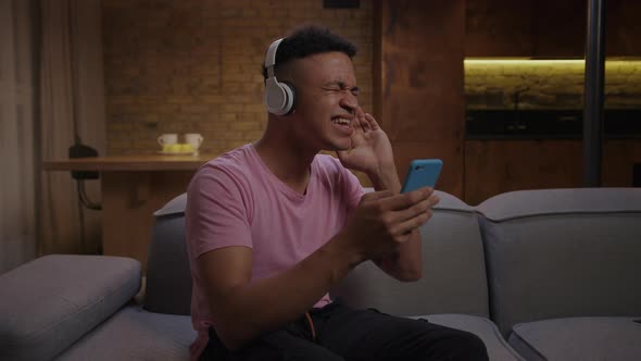 Millennial African American Man in Headphones Singing Karaoke Song Looking at Smartphone Sitting on