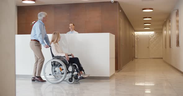 Female Receptionist Greeting Mature Man with Disabled Wife in Wheelchair