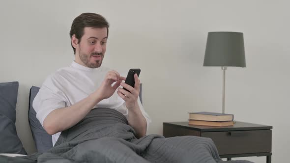 Young Man Celebrating Success on Smartphone in Bed