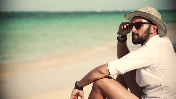 Businessman In Hat Sunbathing Enjoying Ocean Vacation. Man Sitting On Tropical Beach In White Shirt.