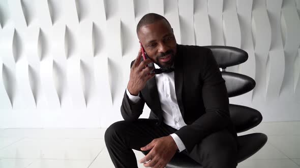 Black Man Dressed Tuxedo Is Talking By Cell Phone Sitting in Studio, Chatting and Smiling