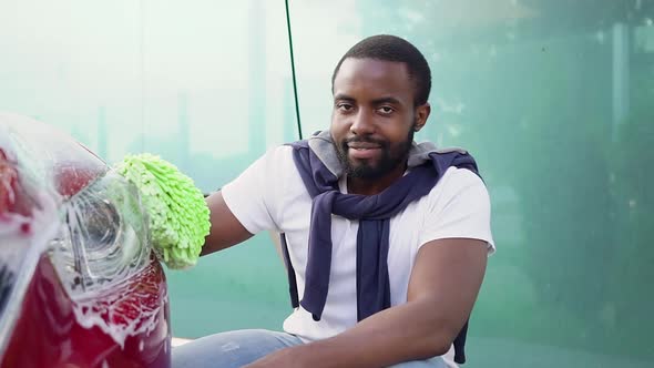 Black-Skinned Guy Looking at Camera while Washing His Car with Hand Glove in Car Wash Station