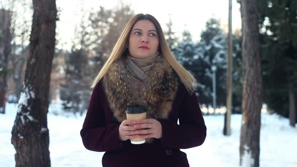 A Beautiful Young Girl is Walking Along the Winter Park and Drinking Coffee