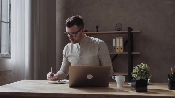 Guy Writing in Notebook While Sitting at Table and Listening To Teacher During Online Lecture at