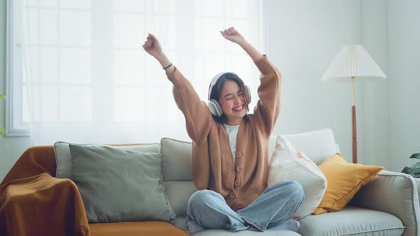 Happy asian woman wear wireless headphones having fun listen music in smartphone and sit on sofa