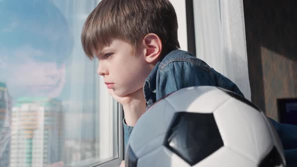 Caucasian Boy on a Home Quarantine Looking Out the Window a Bored Boy Holds a Soccer Ball