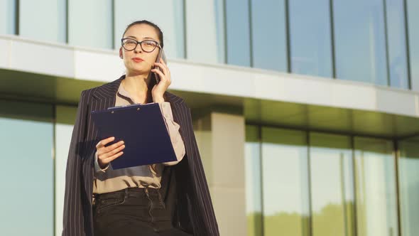 Portrait of businesswoman with glasses talking at the phone, business building in the background