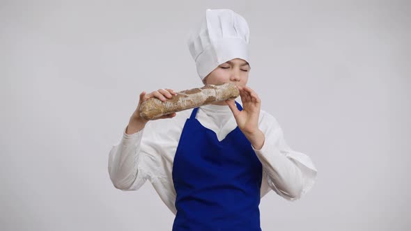 Happy Caucasian Boy in Chef Uniform Enjoying Smell of Freshlybaked Baguette Biting Tasty Food