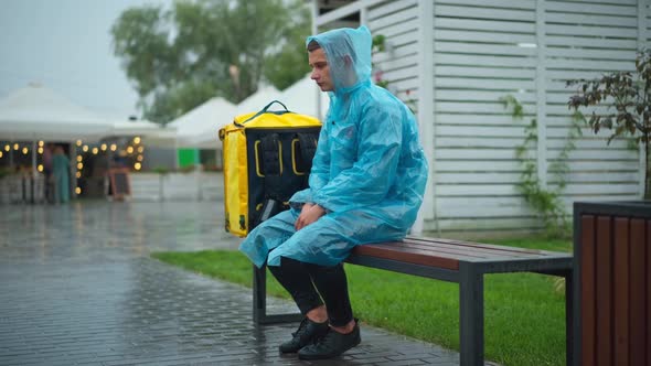 Wide Shot Portrait of Tired Sad Courier in Rain Coat Sitting on Bench in City Sighing Standing Up