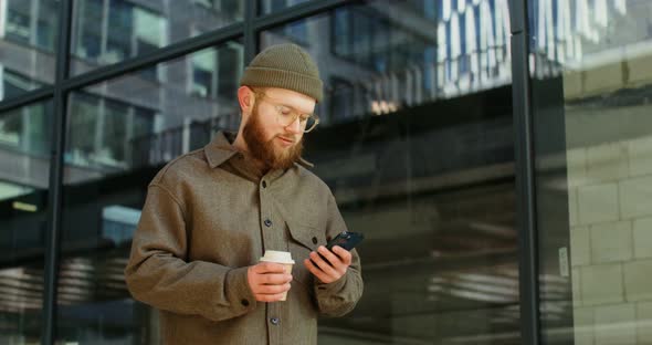 A Bearded Man is Typing on a Mobile Phone While Standing Near Office Building