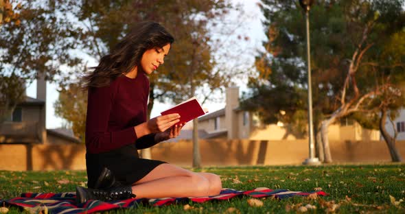 A gorgeous young hispanic woman student reading a book or novel outdoors on a college university cam