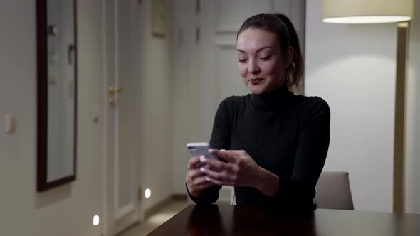 A Brooding Brunette Woman Sits at a Table in the Room and Looks Into Her Smartphone Typing a Text