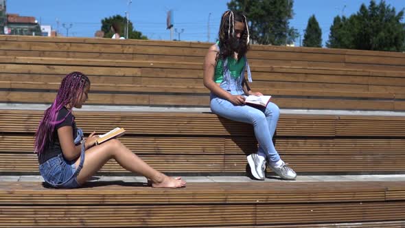 Two Girls are Reading a Book Sitting on a Wooden Bench in the Park in Sunny Weather