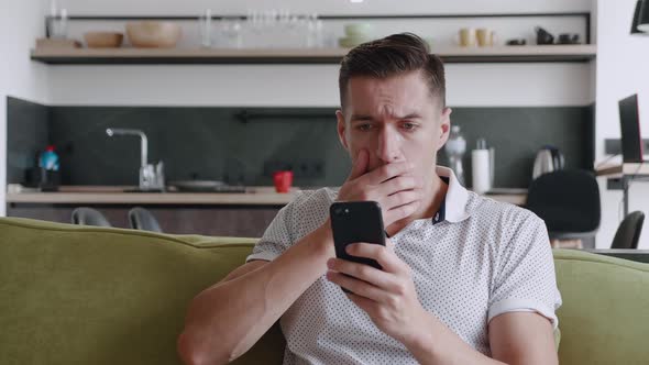 Shocked Man Reacting Message on Smartphone Covers His Mouth in Fright Sitting on Sofa at Home Living