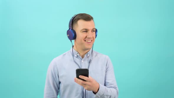 Studio Portrait of Man in a Blue Shirt Chooses Music and Turns It on in Headphones From a Smartphone