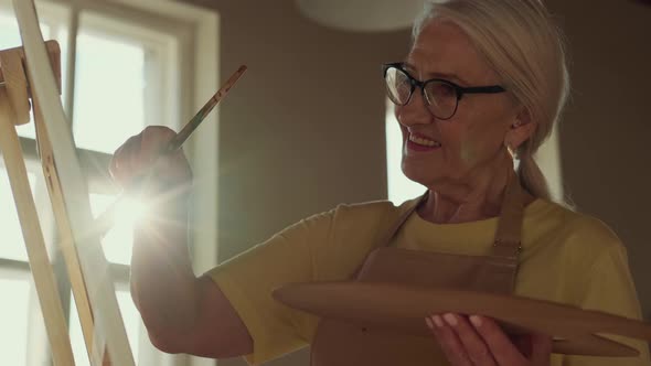 Elderly Woman Artist Paints a Picture on Canvas While Standing in a Sunny Workshop During Sunset