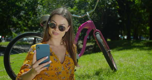 Close-up Beautiful Girl Sitting on the Grass in the Park Near the Bike and Uses a Smartphone