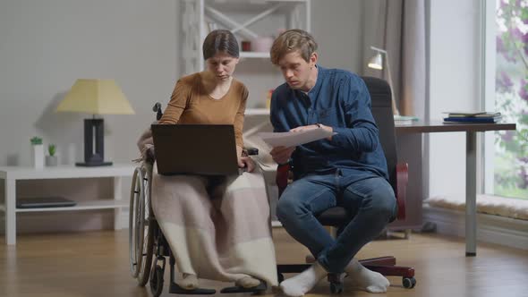 Front View Concentrated Serious Caucasian Woman in Wheelchair and Man Talking Analyzing Paperwork