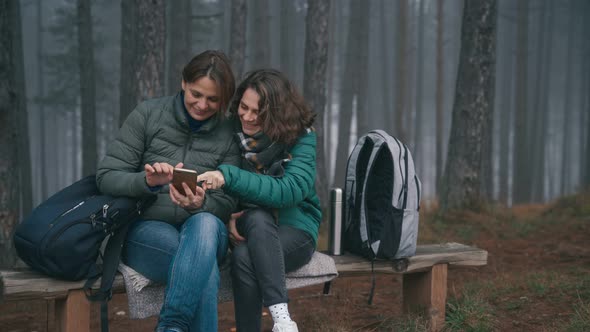 Two Young Women Using a Smartphone While Sitting on a Bench in a Forest 