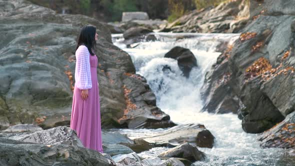 Young pretty woman in long pink fashionable dress standing near small river with fast moving water.