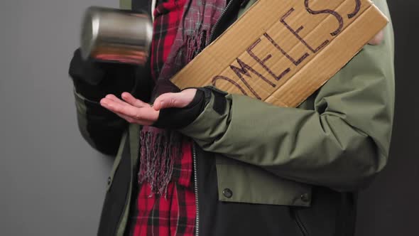 A homeless man in dirty clothes pours coins out of a steel cup and counts them. Close-up