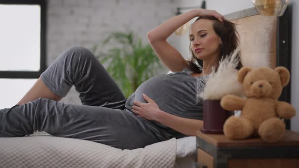 Young Charming Pregnant Woman in Pajamas Lying on Bed Stroking Belly Thinking