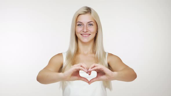 Smiling Caucasian Girl in White with Long Blond Hair Showing Heart Shape By Fingers in Good Mood