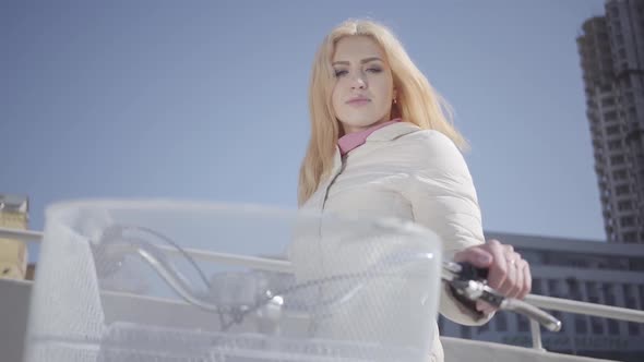 Portrait Cute Young Blond Woman in Warm White Jacket Standing at the City Street with Bicycle