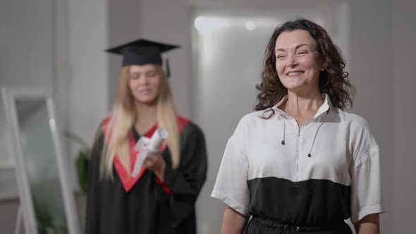 Excited Caucasian Mother Posing at Home Looking Back at Intelligent Graduate Daughter with Rolled