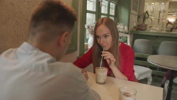 A Man and a Beautiful Young Girl in a Red Dress are Drinking Drinks at a Table in a Cafe and Talking